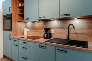 Moderne Küche in einem Junior-Apartment mit hellblauen Schränken, Arbeitsplatten aus Holz, Einbaubackofen, Elektroherd, Spüle mit schwarzer Armatur, Kaffeemaschine und offenen Regalen. Unterbauleuchten sorgen für eine helle Beleuchtung des Arbeitsbereichs.