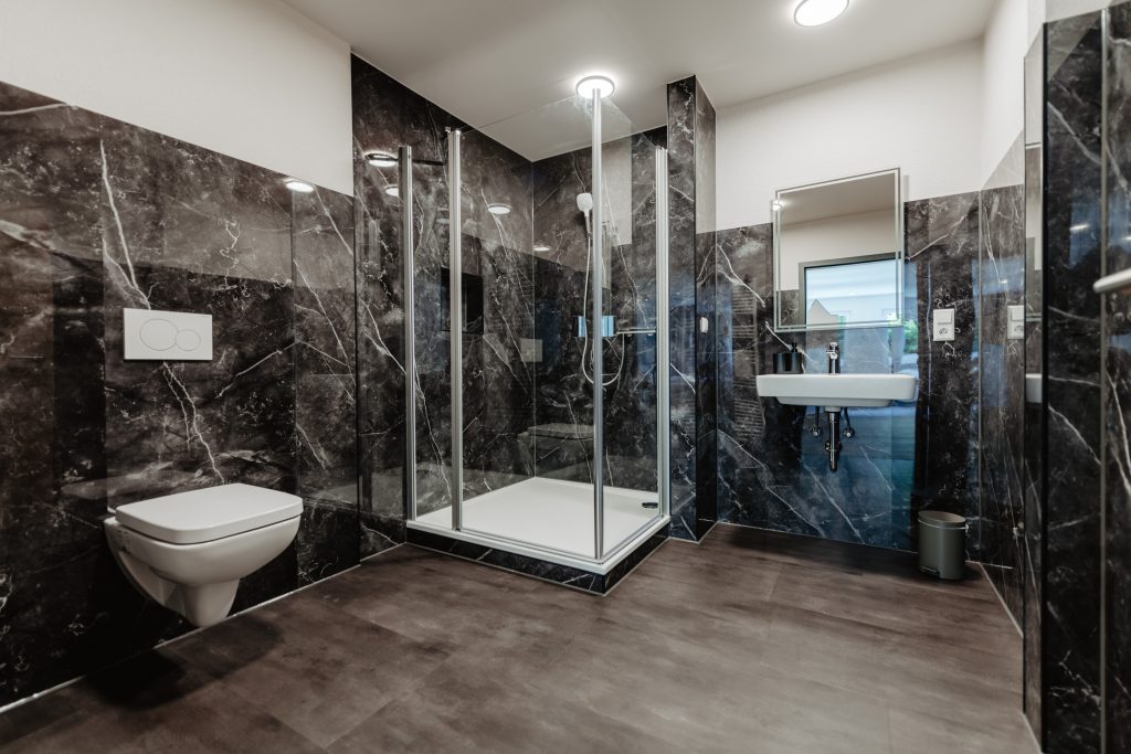 Modernes Badezimmer in einem Junior-Apartment mit dunklen Marmorwänden, einer Duschkabine aus Glas, einem wandhängenden WC, einem wandmontierten Waschbecken mit Spiegel und einem Mülleimer auf dunklem Boden. Der Raum wirkt geräumig und hell.
