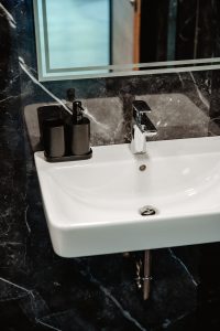 Ein weißes, rechteckiges Waschbecken mit einer verchromten Armatur, einem schwarzen Seifenspender und einem Glas-Set vor einer dunklen Marmorwand unter einem großen Spiegel verleiht diesem Junior-Apartment moderne Eleganz.