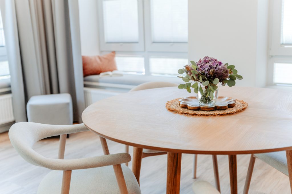 Ein runder Holztisch mit zwei hellen Stühlen, einer Vase mit violetten Blumen und einer geflochtenen Tischunterlage schafft in dieser gemütlichen kleinen Wohnung eine einladende Essecke. Im Hintergrund sind große Fenster und eine Fensterbank mit Kissen zu sehen.