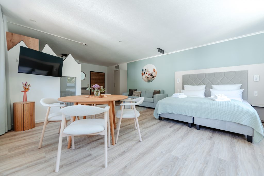 Modernes Zimmer im Junior-Apartment-Hotel mit einem runden Esstisch und vier Stühlen, einem großen Bett mit einer hellblauen Decke, einem an der Wand montierten Fernseher, einem Sofa und einem Wandbild mit Blumenmotiv. Das Zimmer verfügt über Holzböden und ist in sanften Pastelltönen gehalten.