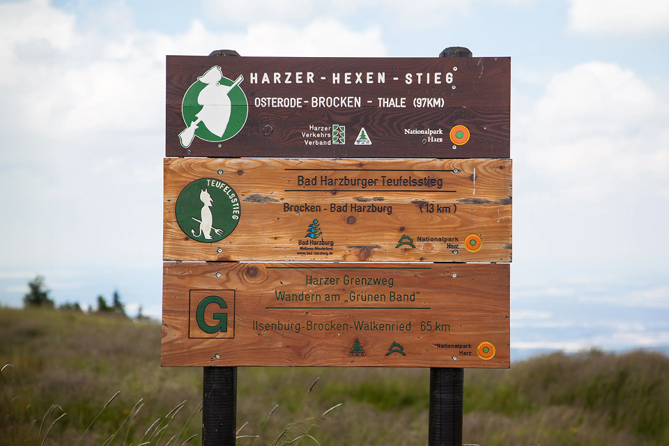 Holzschild für den Wanderweg „Harzer-Hexen-Stieg“ mit Angaben zu Entfernungen und Wegbeschreibungen zu verschiedenen Zielen, mit Logos, einem Hexensymbol und einer grasbewachsenen Landschaft im Hintergrund unter einem bewölkten Himmel.